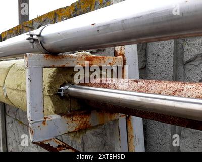 Metal pipes are mounted on a bracket, showcasing rust and insulation material along a concrete fence in an industrial setting. Stock Photo