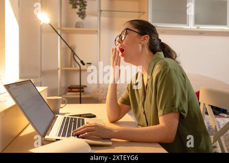 Caucasian young woman is sitting at her desk with a laptop and yawning. Fatigue due to overworking. The student falls asleep near the computer. Online Stock Photo