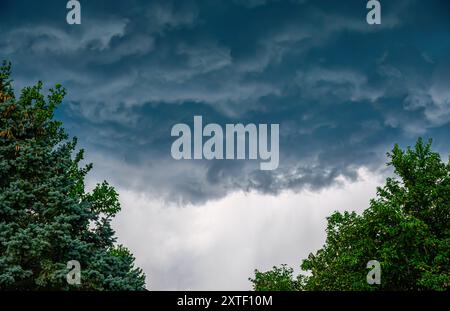 Dark thunderclouds through a cleft of green trees with lighter clouds and precipitation below Stock Photo