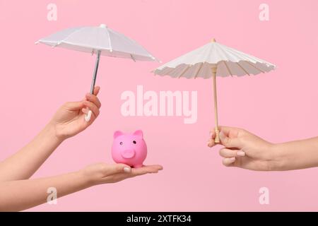 Female hands with mini umbrellas and piggy bank on pink background Stock Photo