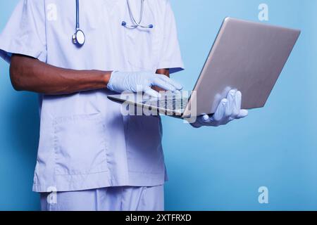 Man working as nurse using wireless computer while wearing stethoscope, gloves for protection and scrubs. African american individual standing and utilizing a digital laptop. Stock Photo