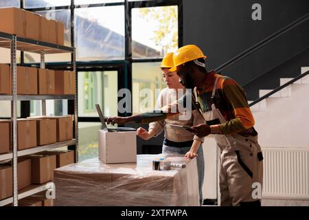 Warehouse employees preparing clients orders using cardboard boxes, checking delivery details on laptop computer in storage room. Diverse colleagues working at products inventory report in storehouse Stock Photo
