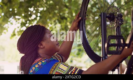 Young african american girlfriend carrying wheel and positioning it on bicycle body with her caucasian boyfriend. Outdoors multiracial couple doing bike tire maintenance. Close-up side-view shot. Stock Photo