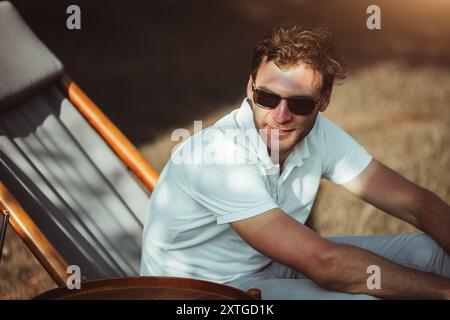 A man wearing sunglasses sits in a wooden deck chair, enjoying a sunny day outdoors. He is dressed casually in a white polo shirt and light pants, smi Stock Photo