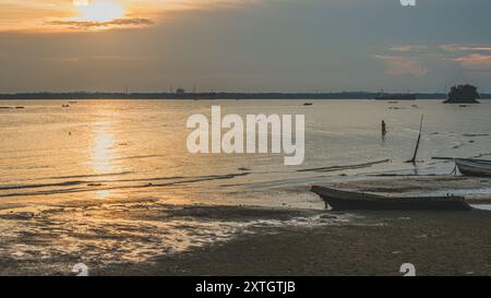 Balikpapan, Indonesia - June 15th, 2024. A peaceful scene of a colorful sunset over a calm beach with a wooden boat resting on the shore. Stock Photo