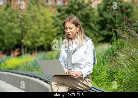 Focused woman working online remotely via internet in public park sitting on bench using laptop. Stock Photo