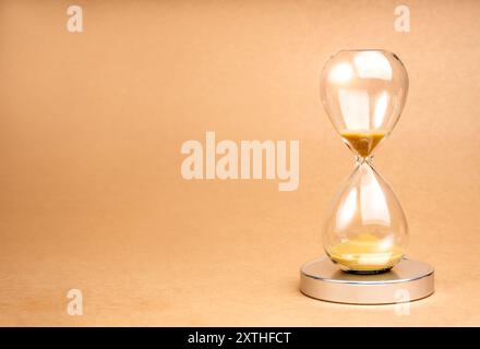 A clear hourglass with gold sand on chrome podium on light brown recycled paper background with copy space. Golden sand in sandglass clock isolated on Stock Photo