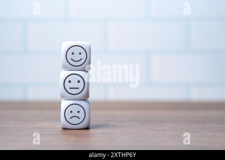 Three white dice with happy, neutral and sad face are stacked on a wooden surface in front of a white brick wall. Copy space for text. Emotion, mood a Stock Photo