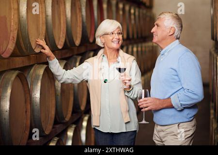 Wine cellar, barrel and senior couple with glass, talking and celebration with alcohol, industry and retirement. Happiness, woman and man with Stock Photo