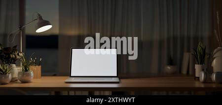 A laptop computer with a white-screen mockup on a wooden table by the window in a house at night, illuminated by a table lamp. 3d render, 3d illustrat Stock Photo
