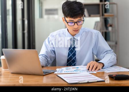 Young businessman working on his project in his office checking business result data from the printed sheets and his laptop computer Stock Photo