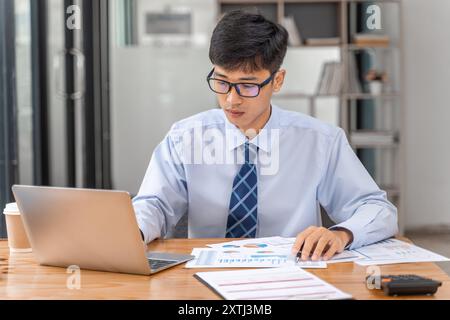Young businessman working on his project in his office checking business result data from the printed sheets and his laptop computer Stock Photo