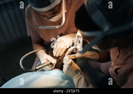 Close-up of two female doctors in lighting glasses extracting hair follicles from donor area of male patient for hair transplantation in clinic. Stock Photo