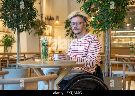 Alone thoughtful disabled young man on wheelchair spending time in cafe drinking coffee reflects.  Stock Photo