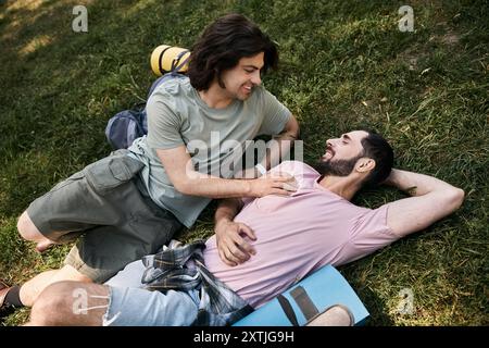 Two young men, a gay couple, enjoy a summer hike, laying on the grass and smiling at each other. Stock Photo