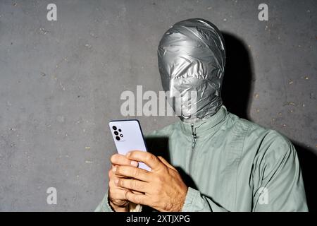 Mystery man with duct tape face types text message on cell phone, enjoys online communication on gray background Stock Photo