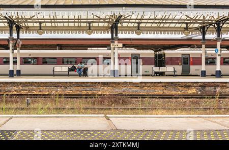 A modern train stands at a historic railway station platform under a 19th Century iron canopy. A man sits on a bench with his suitcase. Stock Photo