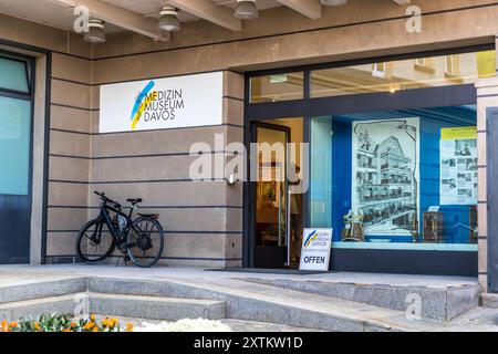 The Davos Medicine Museum shows, on its façade on the Platzstrasse, Grisons, a logo with a stylised fever thermometer and an 'Offen' (Open) sign beside the entrance. Historic black-and-white photographs and information panels are visible in the illuminated shop window, and a bicycle leans against the façade, Switzerland. Stock Photo