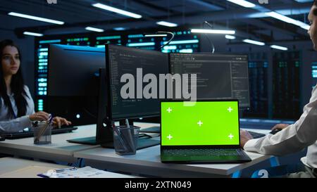 Zoom in on green screen laptop used by data center worker troubleshooting issues. Engineer uses chroma key notebook in server hub, establishing performance benchmarks, camera B Stock Photo