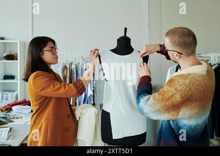 Two young fashion designers standing in front of mannequin and putting unfinished dress on dummy while working over new collection Stock Photo