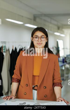 Young confident tailor in smart casualwear looking at camera while standing by workplace with sketches of new attire Stock Photo