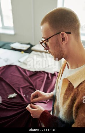 Focus on young man in eyeglasses and stylish attire standing in front of camera and stitching parts of purple textile together Stock Photo