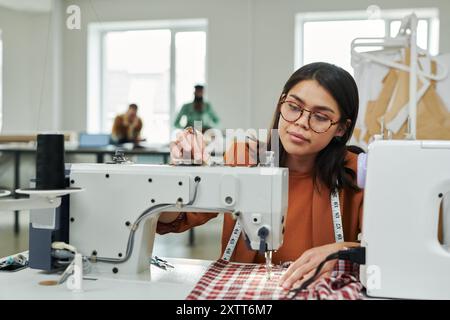 Young female tailor adjusting electric sewing machine before stitching while sitting by workplace against two coworkers in atelier Stock Photo