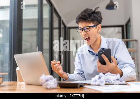 Frustrated and angry young businessman working late at the office trying to finish his project holding a phone in one hand looking angry at the screen Stock Photo