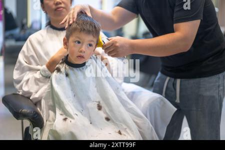 In a bustling salon, a young boy receives a trim while being securely held by a caretaker, keeping him calm and still during his haircut. Stock Photo