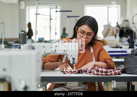 Young female fashion designer or seamstress stitching piece of checkered fabric while sitting by electric sewing machine in atelier Stock Photo