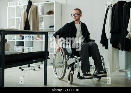 Young male fashion designer with disability moving along spacious workshop or atelier in wheelchair during working day Stock Photo