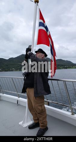 A senior man takes photos with his smartphone during a fjord tour in Norway. Behind him the Norwegian flag Stock Photo