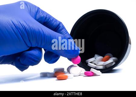 Blue gloved doctor's hand holding pink color pill from multicolored pill spilled from black container on white background. Stock Photo