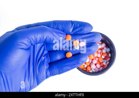 Upward view of a hand wearing blue gloves holding pink and orange pills, with lots of pills in a black container on a white background. Stock Photo