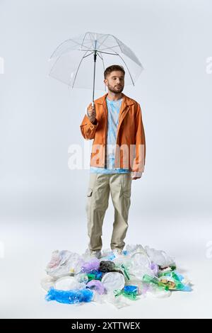 A man with an orange jacket and a clear umbrella stands above a large pile of plastic waste. Stock Photo