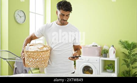A handsome young hispanic man multitasks with laundry and a smartphone in a home interior. Stock Photo