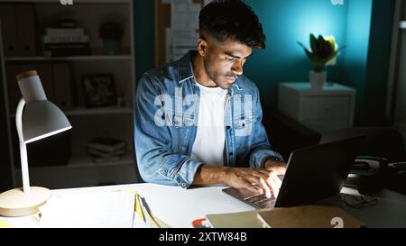 A focused hispanic man with a beard works on a laptop in a modern office room, illuminated by a desk lamp at night. Stock Photo