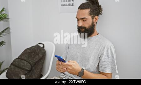 A bearded young hispanic man sits in a modern indoor waiting room, focusing on a smartphone beside a backpack. Stock Photo