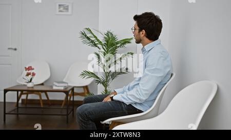 A bearded young man in a blue shirt sits contemplatively in a modernly furnished white room with plant decor. Stock Photo