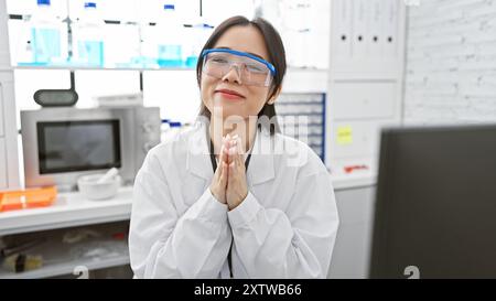 A young asian woman smiles in a laboratory setting, wearing safety glasses and a lab coat, suggesting a professional scientific environment. Stock Photo