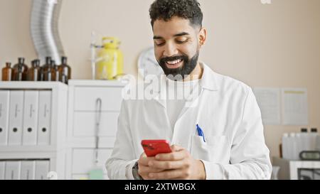 Handsome hispanic male lab technician in white coat using smartphone in pharmacy Stock Photo