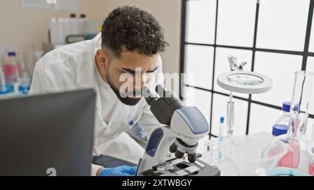 A focused hispanic man with a beard examines samples using a microscope in a modern laboratory setting. Stock Photo