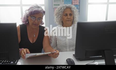 Women collaborating maturely in an office setting, both focused on a business document while seated at desks with computers nearby, highlighting teamw Stock Photo