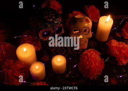 A vivid Día de los Muertos scene featuring two skulls: one adorned with metallic flowers and another hollow with a lit candle inside. Surrounded by ma Stock Photo