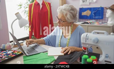Senior woman tailoring in a well-equipped atelier, using a laptop by a sewing machine and colorful fabrics. Stock Photo