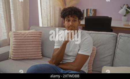 Young man sitting on a couch in a cozy living room, contemplating deeply with a thoughtful expression in a modern apartment setting. Stock Photo