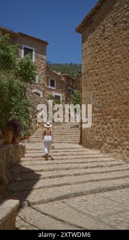 Woman walking on stone steps in fornalutx on a sunny day, showcasing the rustic charm of mallorca, spain with lush greenery and charming stone houses. Stock Photo