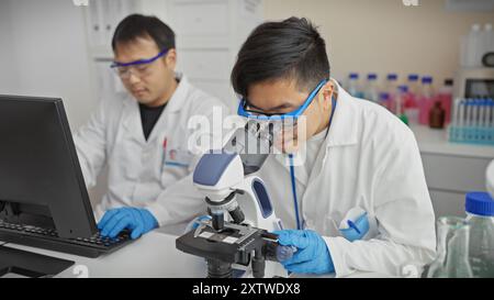 Two men working collaboratively in a modern laboratory, one using a microscope and another on a computer. Stock Photo