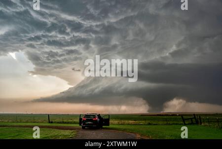 A storm chaser's vehicle parked on a dirt road observing a massive tornado forming under a dark, dramatic sky in an open field. Stock Photo