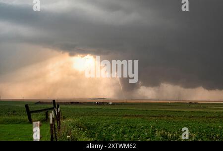 A tornado touches down in a rural area under a stormy sky, with sunlight breaking through clouds. Stock Photo